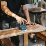 A craftsman uses a power sander on a wooden plank in a well-lit workshop.