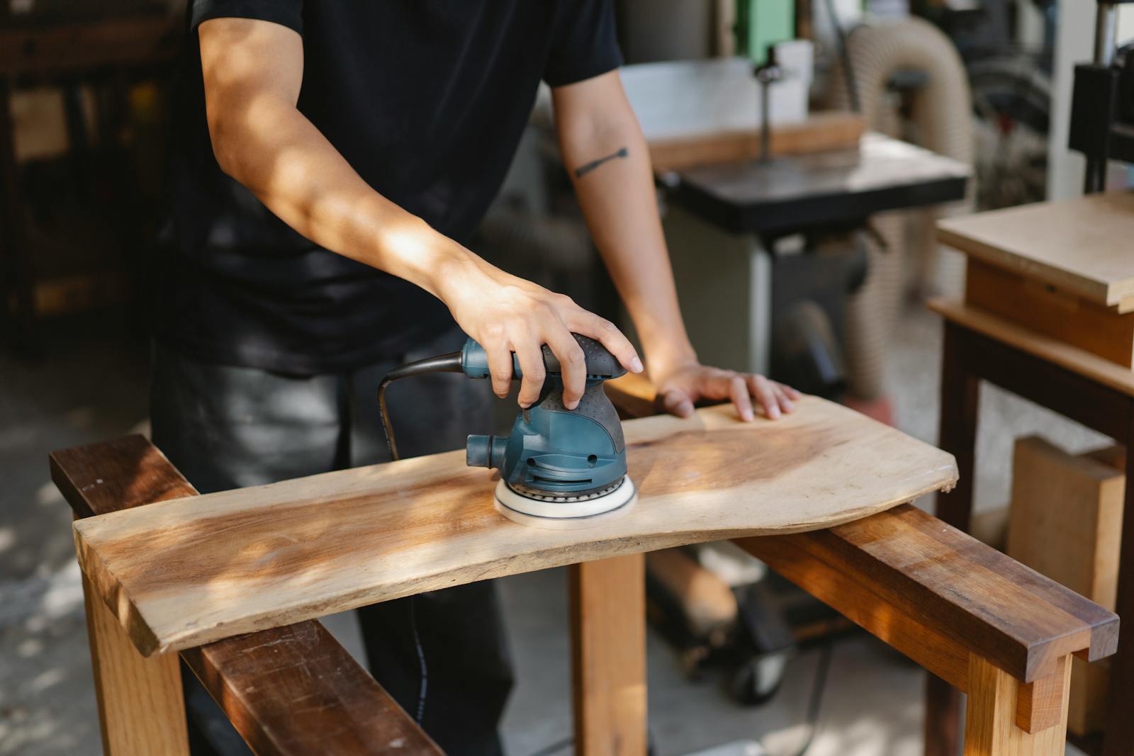 A craftsman uses a power sander on a wooden plank in a well-lit workshop.